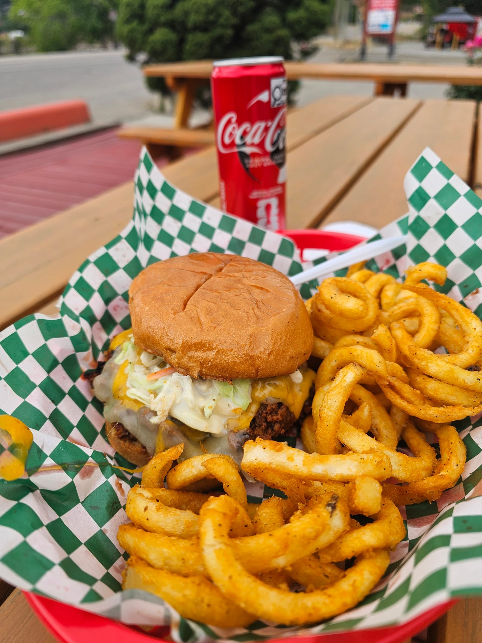 A Deluxe Joe with melted cheese and coleslaw beside curly fries in a checkered-paper basket with a can of Coke, on a picnic table at Lu Lou's in Oyama