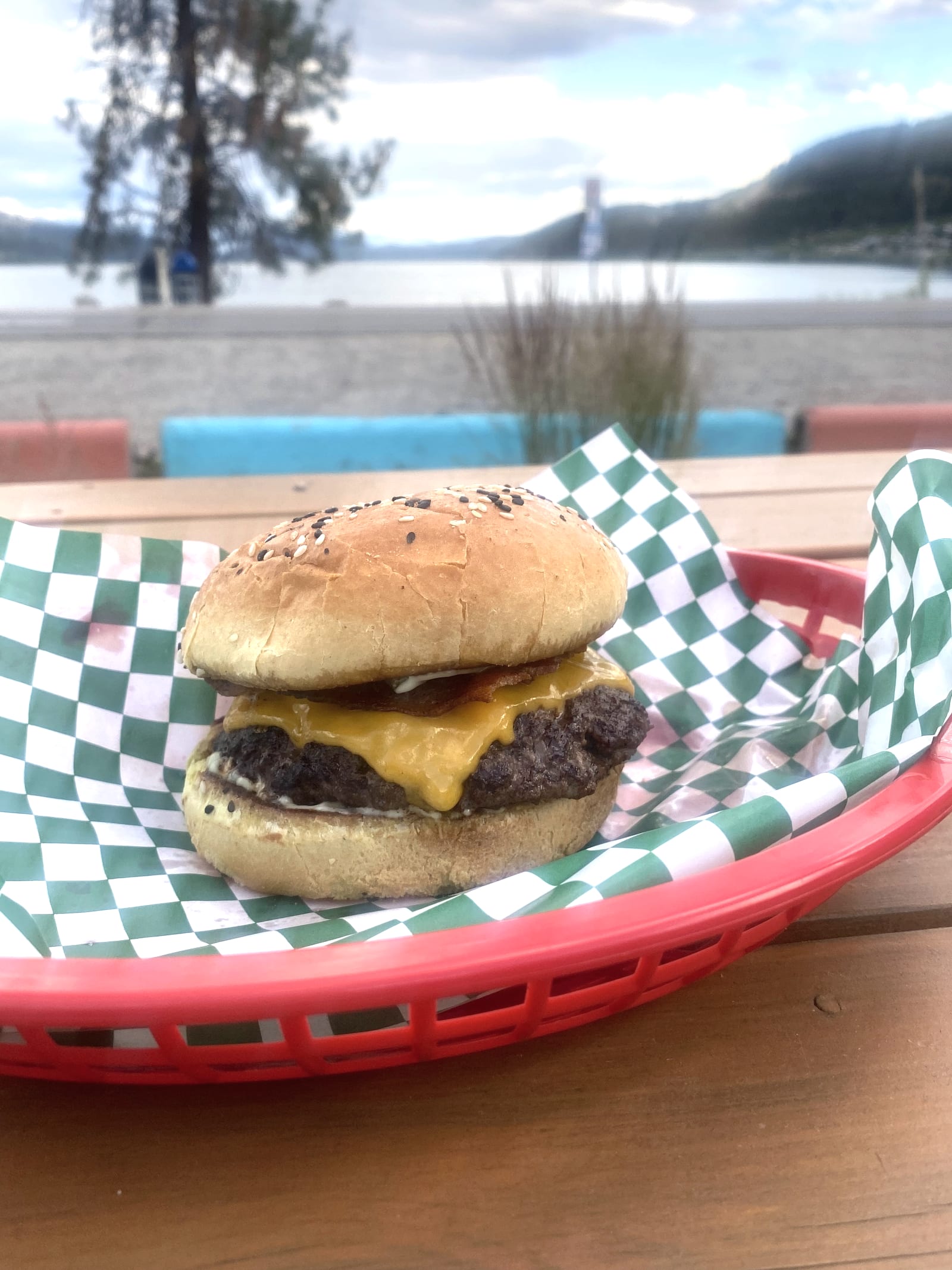 A Lu Lou's smash burger, seared smash patty with melted cheddar and bacon on a toasted sesame bun, in a checker-lined basket with Wood Lake behind